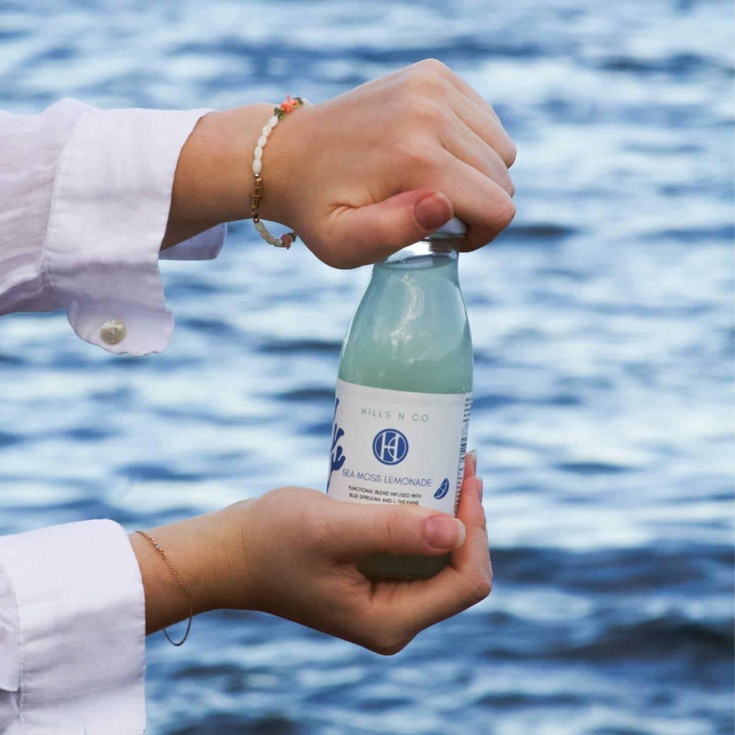 Hills N Co Blue Spirulina Sea Moss Lemonade bottle held by hands with bracelets, against a blue water background.