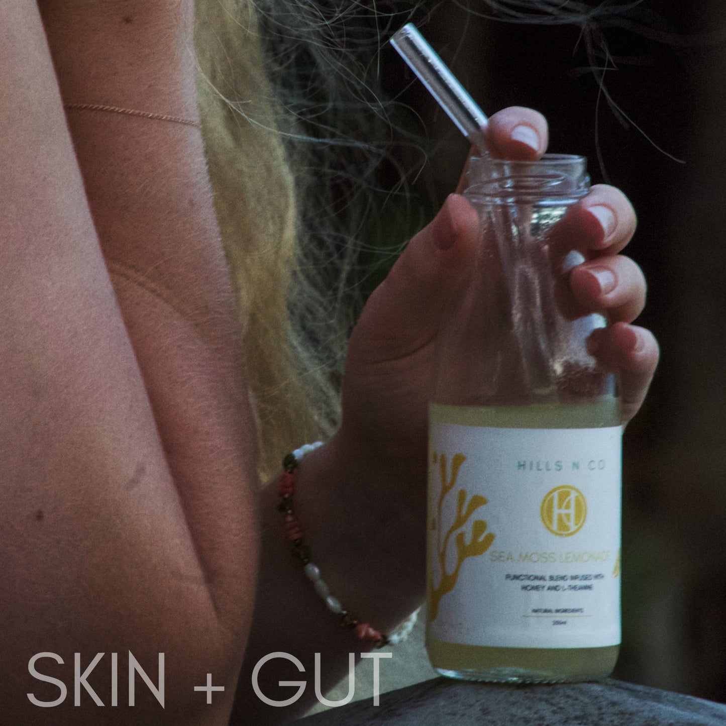 Hills N Co Honey Sea Moss Lemonade bottle with a glass straw, held by a woman with blonde hair and a bracelet.